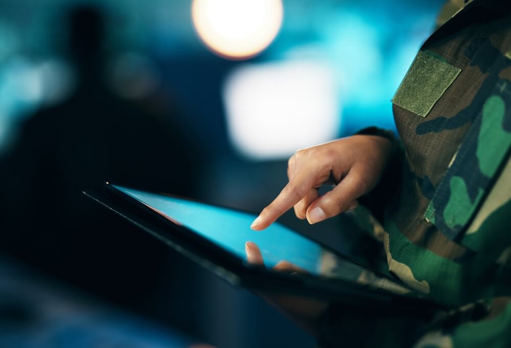 A close-up of a veteran in military uniform using a tablet. The background is blurred, with a bright circular light in the distance. The individual is focusing on the screen, and only part of their uniform and hand are visible.